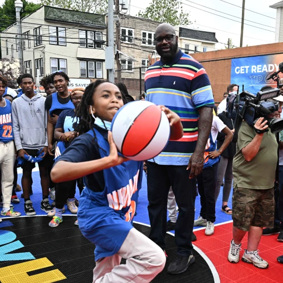 A young girl playing with a basket ball at one of the Newark Comebaq Court, while Shaquille O'Neil and several other people stand in the background.
